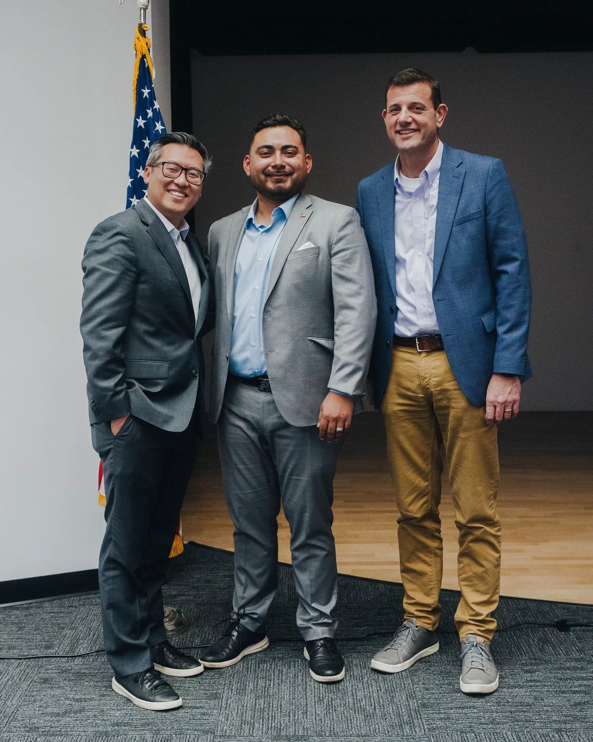 Three men in jackets standing together and smiling in front of an American flag.
