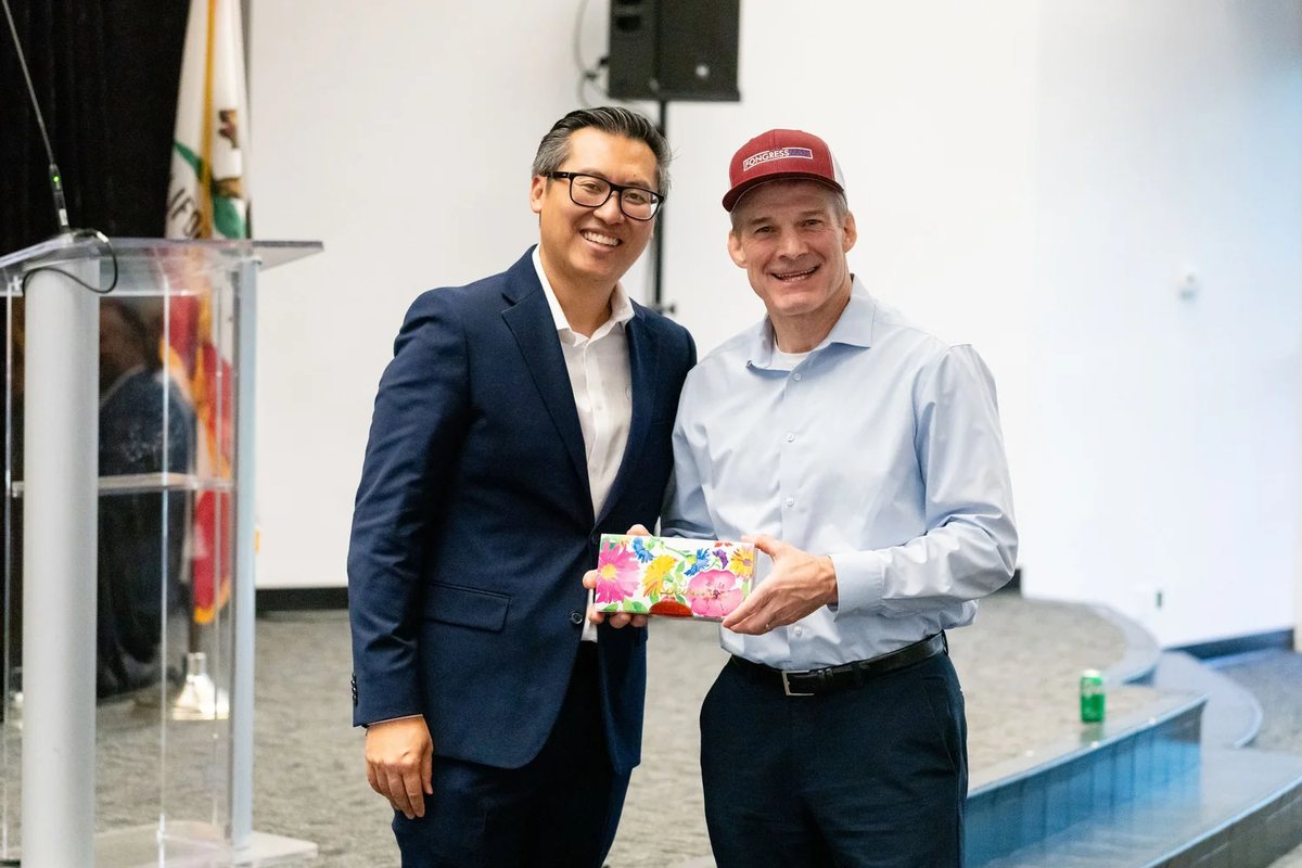 Two men smiling beside a clear podium at the dinner, holding a gift together.