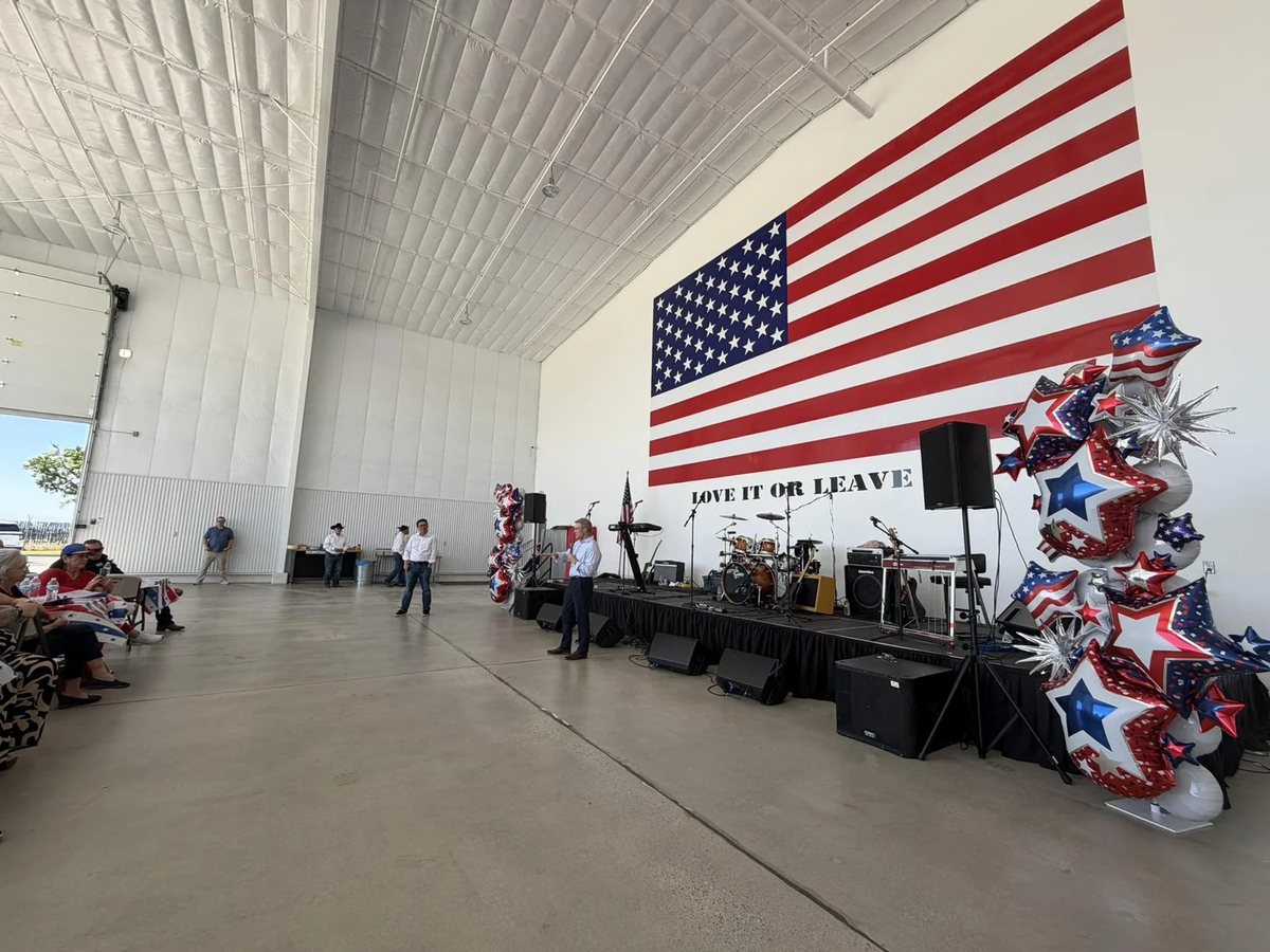 A hangar set for the rally: a large American flag on the wall above a stage with a band's instruments, and red, white and blue star balloons.